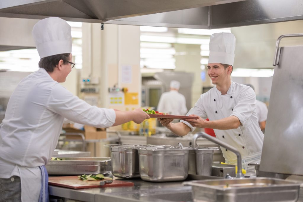 Ein Azubi in der Küche des Klinikums Ingolstadt übergibt einem anderen Azubi ein Tablett.