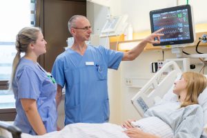 Ein Praxisanleiter zeigt einer jungen Pflegekraft wie sie die Vitalwerte einer Patientin am Bildschirm richtig abliest.