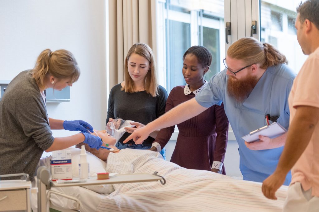 Eine Pflegefachkraft zeigt drei Auszubildenden wie man die Region um einen Katheter am Hals richtig säubert.